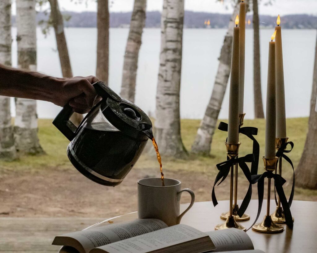 pouring coffee on table with book and candles in front of wintry lake scene