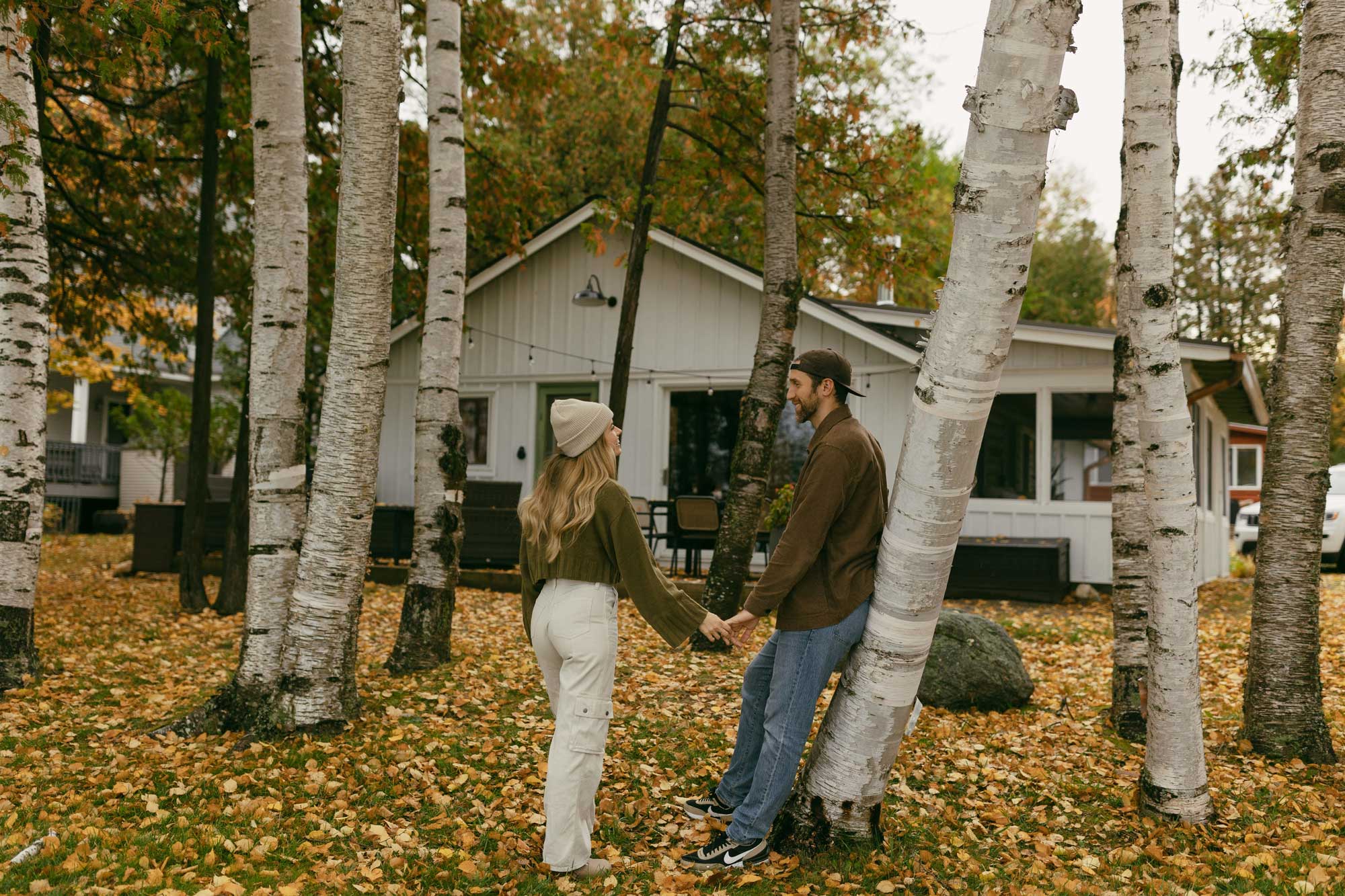 man and woman holding hands standing among birch trees in front of white cabin during fall